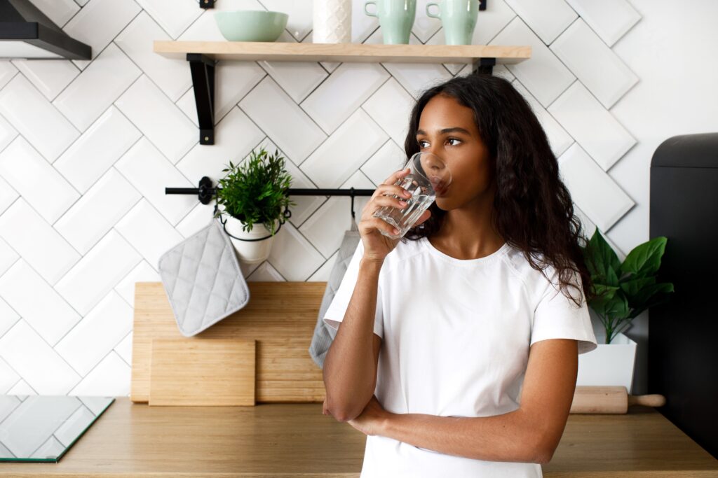 Woman with long curly hair drinking a glass of water in a modern kitchen setting.