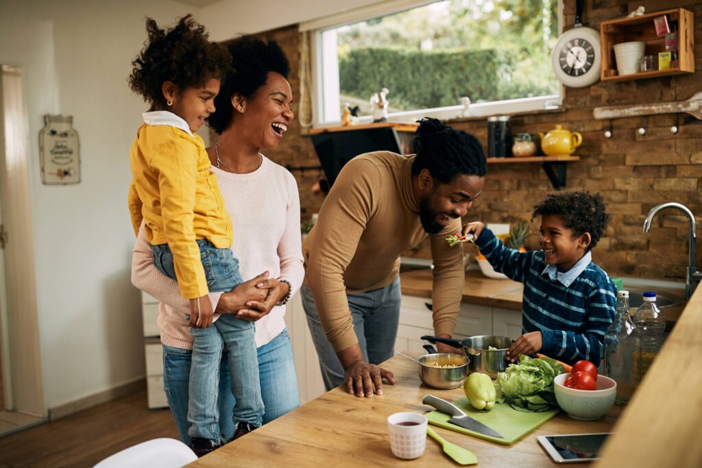 A family of four enjoying cooking together in a kitchen, with one child being held and another preparing food.