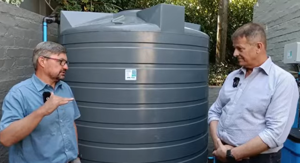 Two men engaged in conversation next to a large gray water storage tank in an outdoor setting.