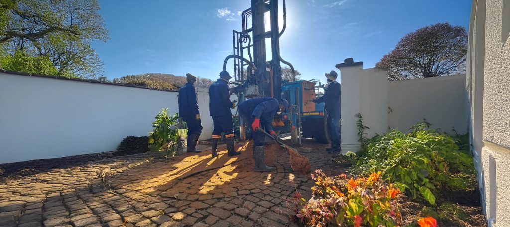 Workers operating a borehole drilling machine in a residential area with clear blue sky.