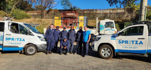 Spritza borehole drilling team in blue uniforms standing beside white Spritza vehicles and a drilling rig, with work site equipment outdoors.