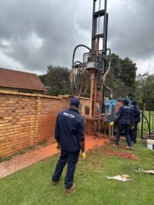 On a borehole drilling site, Spritza technicians in blue uniforms operate a drilling rig beside a brick wall under an overcast sky.