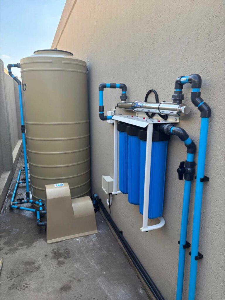 Spritza borehole solutions hardware next to a concrete-grey wall, with blue pipework, valves and cylindrical filtration tanks supporting Johannesburg winter water needs.